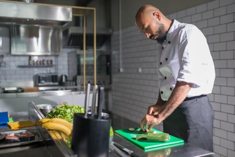 A professional chef in a white jacket uses the claw grip to slice fresh vegetables on a green cutting board in a modern kitchen.