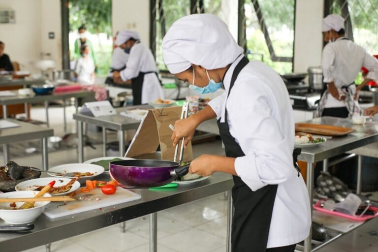 Female culinary student in white chef uniform holding stainless steel tongs in a professional training kitchen.