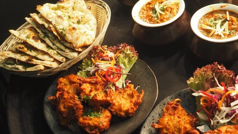 A vibrant spread of traditional Indian vegetarian dishes including curries, flatbreads, and fried snacks arranged on a dark surface.