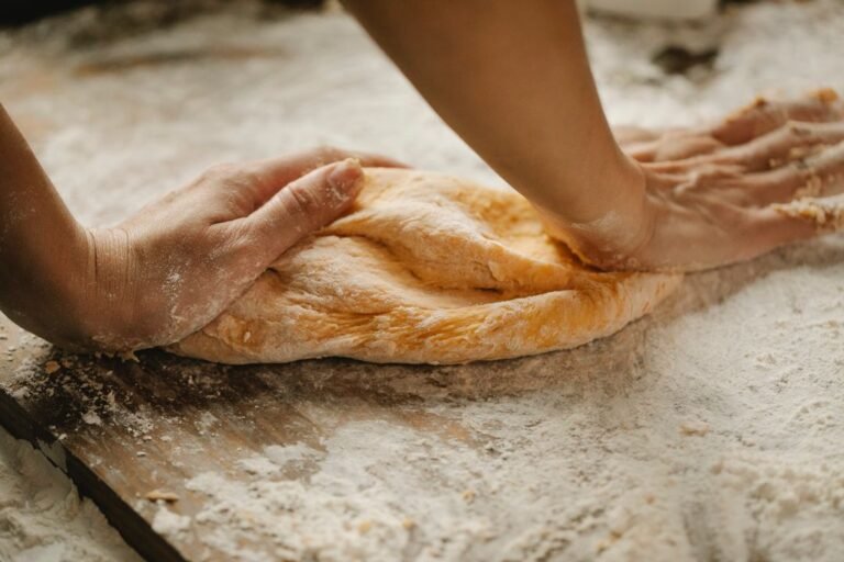 A professional pastry chef kneads fresh dough on a wooden table in a commercial bakery kitchen.