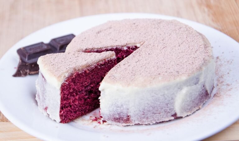 A sliced red velvet cake with cream cheese frosting revealing moist crimson layers inside on a white plate.