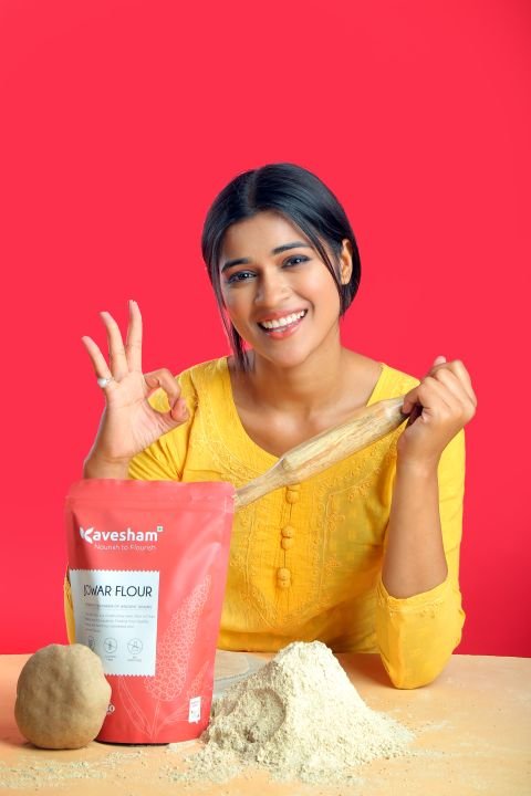 A South Asian woman joyfully baking in a bright kitchen, holding a rolling pin and surrounded by flour and baking ingredients.