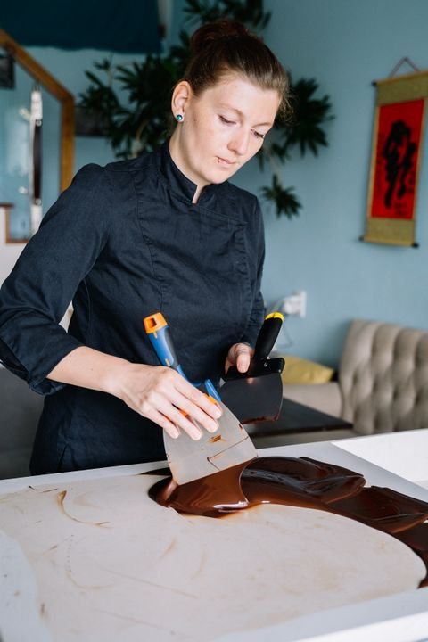 A pastry chef spreads tempered dark couverture chocolate on a marble surface using a metal scraper in a professional kitchen.