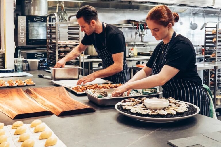 Two professional chefs in white uniforms collaborating over food preparation in a modern hotel kitchen.