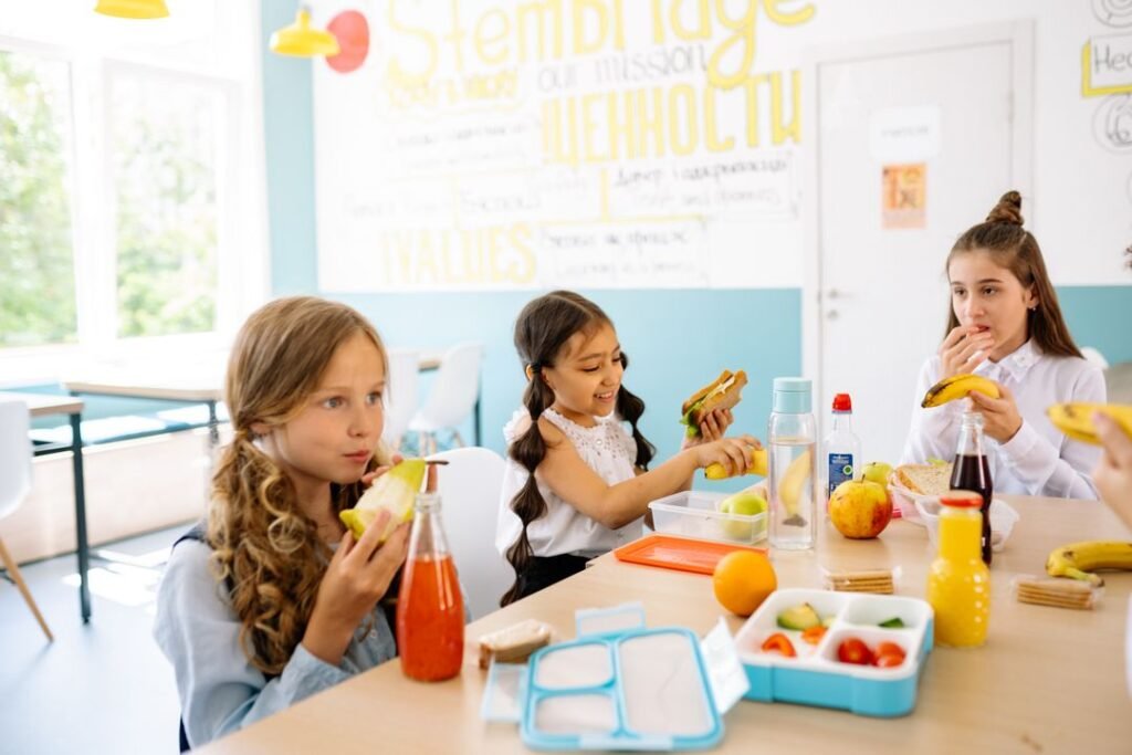 Schoolchildren sharing a healthy packed lunch of fruit and vegetables at a canteen table.