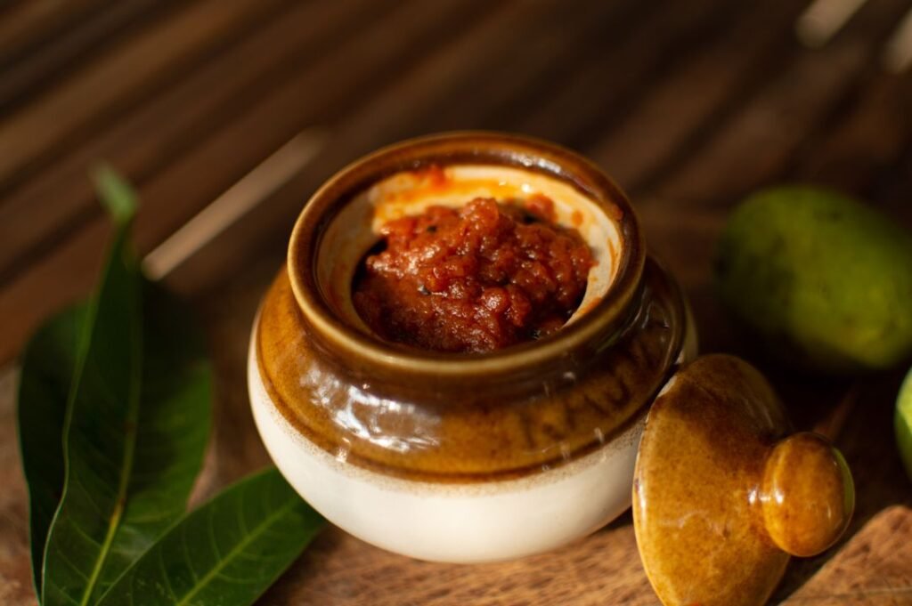 A traditional glazed ceramic pickle jar filled with red mango achaar, surrounded by fresh mango leaves and a whole green mango on a wooden surface.