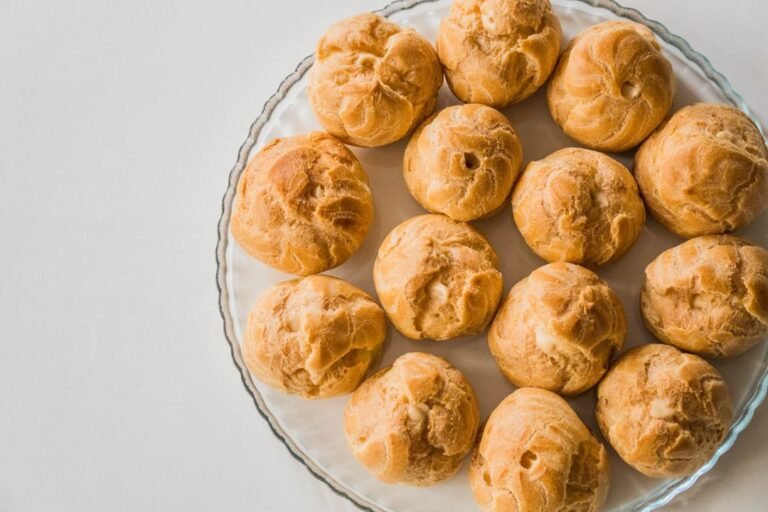 A tray of golden baked cream puffs arranged on a glass plate, the classic result of well-made choux pastry.