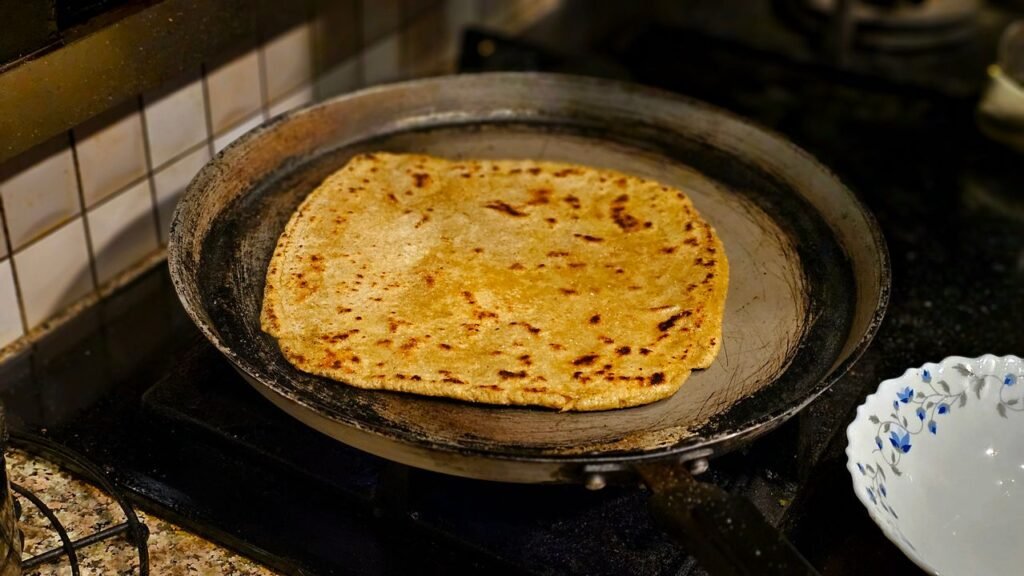 A thick rustic Indian flatbread cooking on a hot pan, showing the coarse texture of a hand-patted rotla.