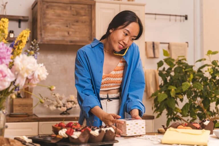 A home baker packing cupcakes into a box while taking a customer order on the phone.
