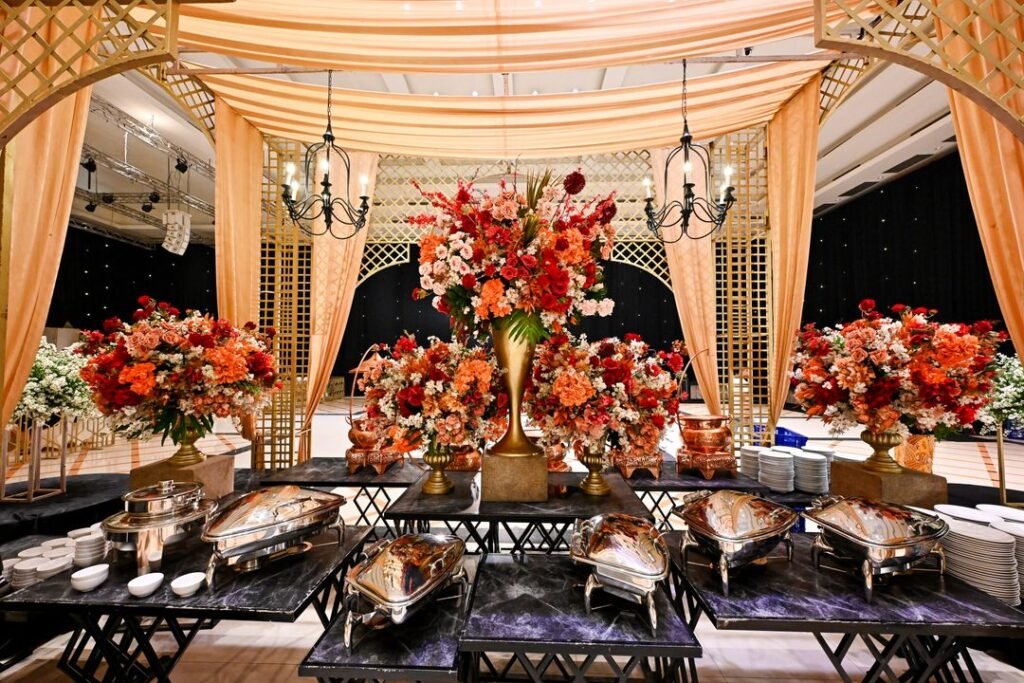 A wedding banquet setup with polished chafing dishes, stacked service plates and large floral arrangements under draped canopies.