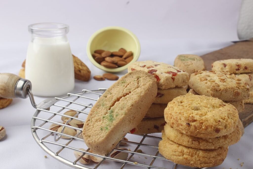 Eggless Nankhatai for Diwali: A Published Base Formula, Variations and Gifting Tips 3 Stacks of nut-topped and tutti-frutti shortbread-style cookies on a steel cooling rack beside a jar of milk and a bowl of almonds.